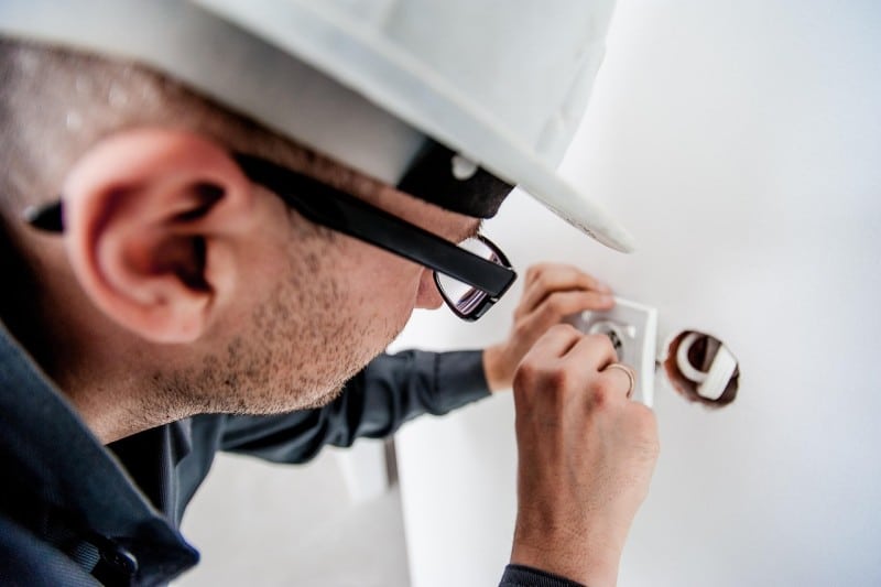 Electrician wearing safety glasses and a hard hat fixing a wall power outlet with a screwdriver, showing simple maintenance to help homeowners avoid costly emergency repair bills.