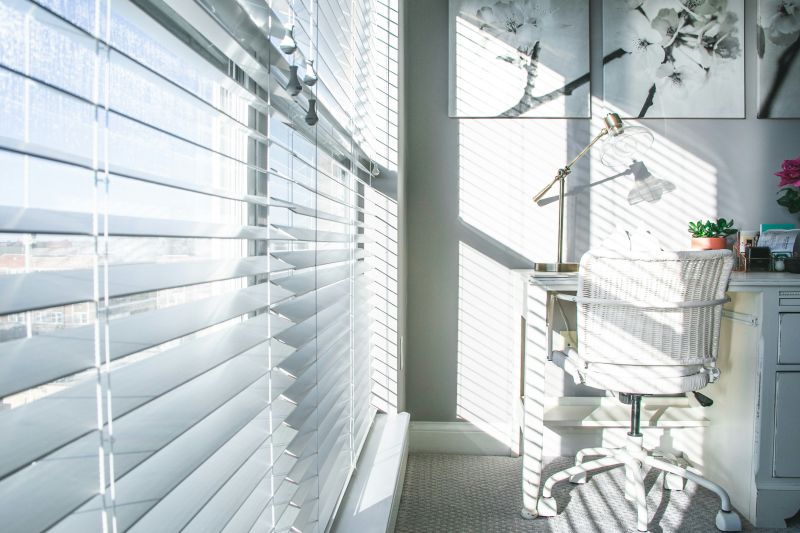 Bright home office with large windows and white blinds allowing natural light to filter in, casting soft shadows on the desk and chair.