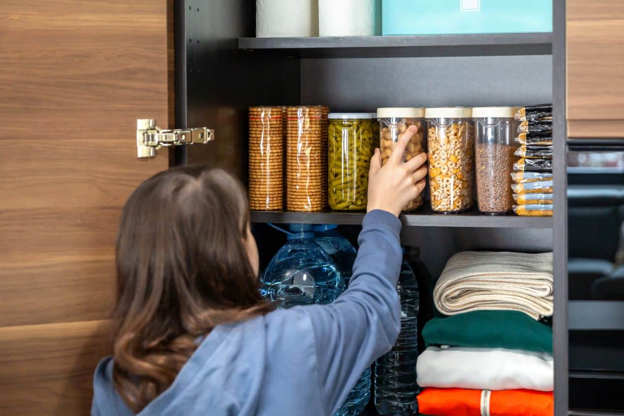 Woman reaching for a food container in a neatly organized pantry stocked with snacks, grains, bottled water, and folded linens.