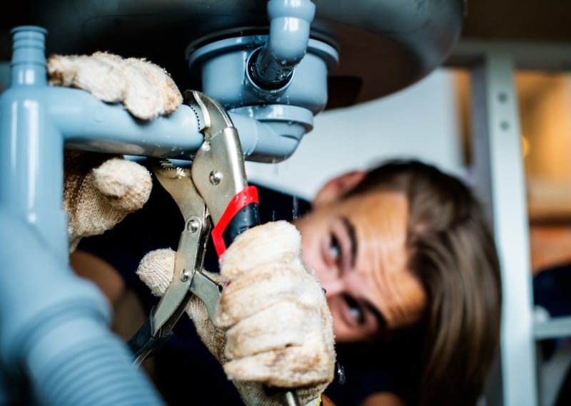 Plumber tightening sink drain pipe with pliers, wearing gloves, performing plumbing maintenance to fix leaks and ensure system reliability.