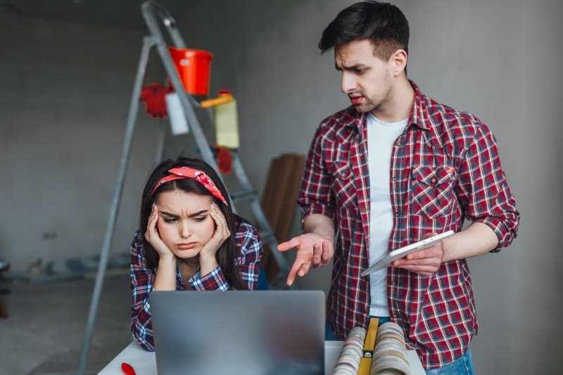 A frustrated woman sits at a laptop holding her head while a man beside her gestures with a tablet, both wearing plaid shirts, in a room under renovation. The scene illustrates common remodeling mistakes homeowners regret.