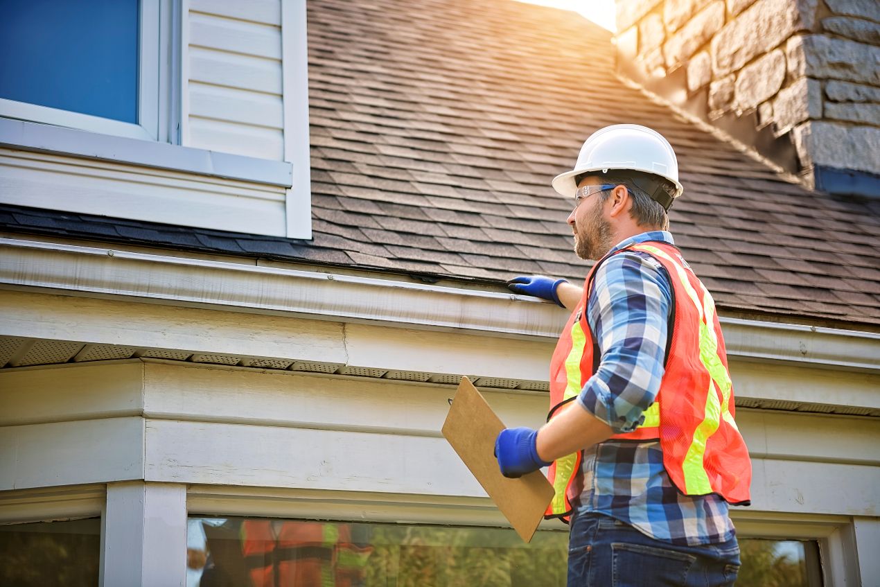 A roofing inspector in safety gear examining roof shingles and gutters to assess whether repair or replacement is needed.