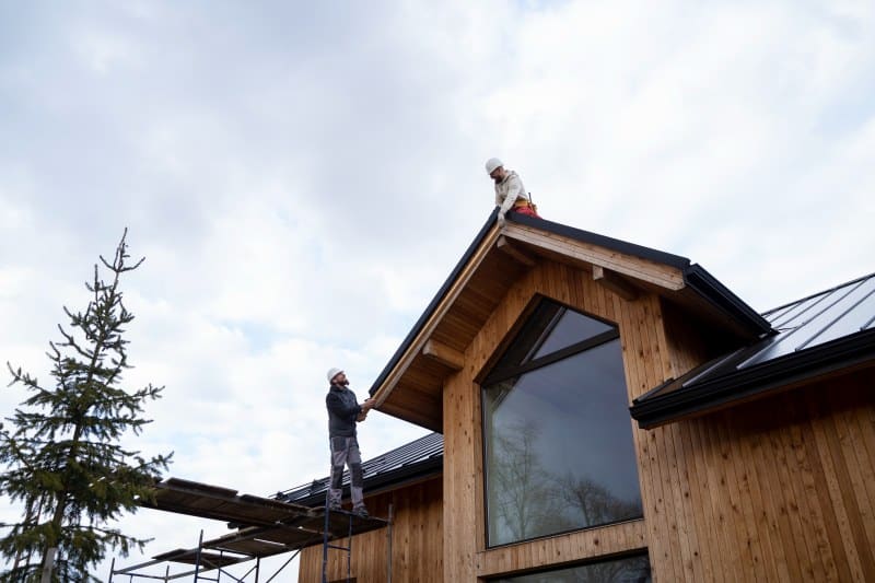 A construction worker in safety gear repairs a damaged roof, standing on a ladder placed against red roof tiles.