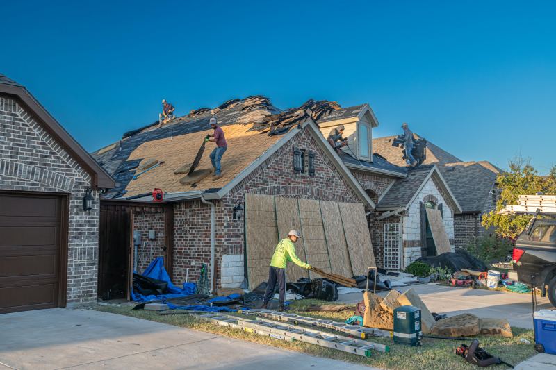 Workers replacing shingles on a brick house during emergency roof repair, with tools and materials scattered around the driveway.