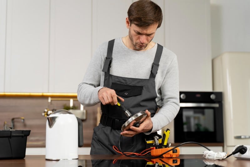 A repair technician in overalls uses a screwdriver to maintain a kitchen appliance part, with tools and an electric kettle nearby.