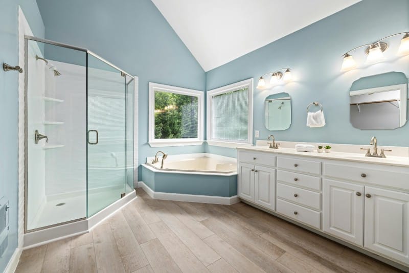Spacious bathroom with a glass-enclosed shower, corner bathtub under large windows, dual sinks with mirrors, and white cabinets. Light blue walls and wood-style flooring create a relaxing spa bathroom feel.