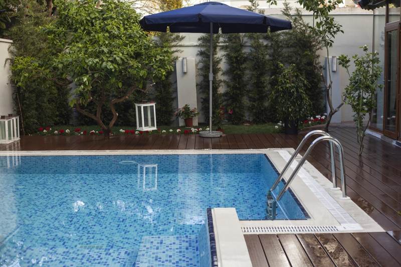 Outdoor swimming pool with mosaic blue tiles, stainless steel ladder, wooden deck, umbrella, and surrounding greenery including trees and plants.