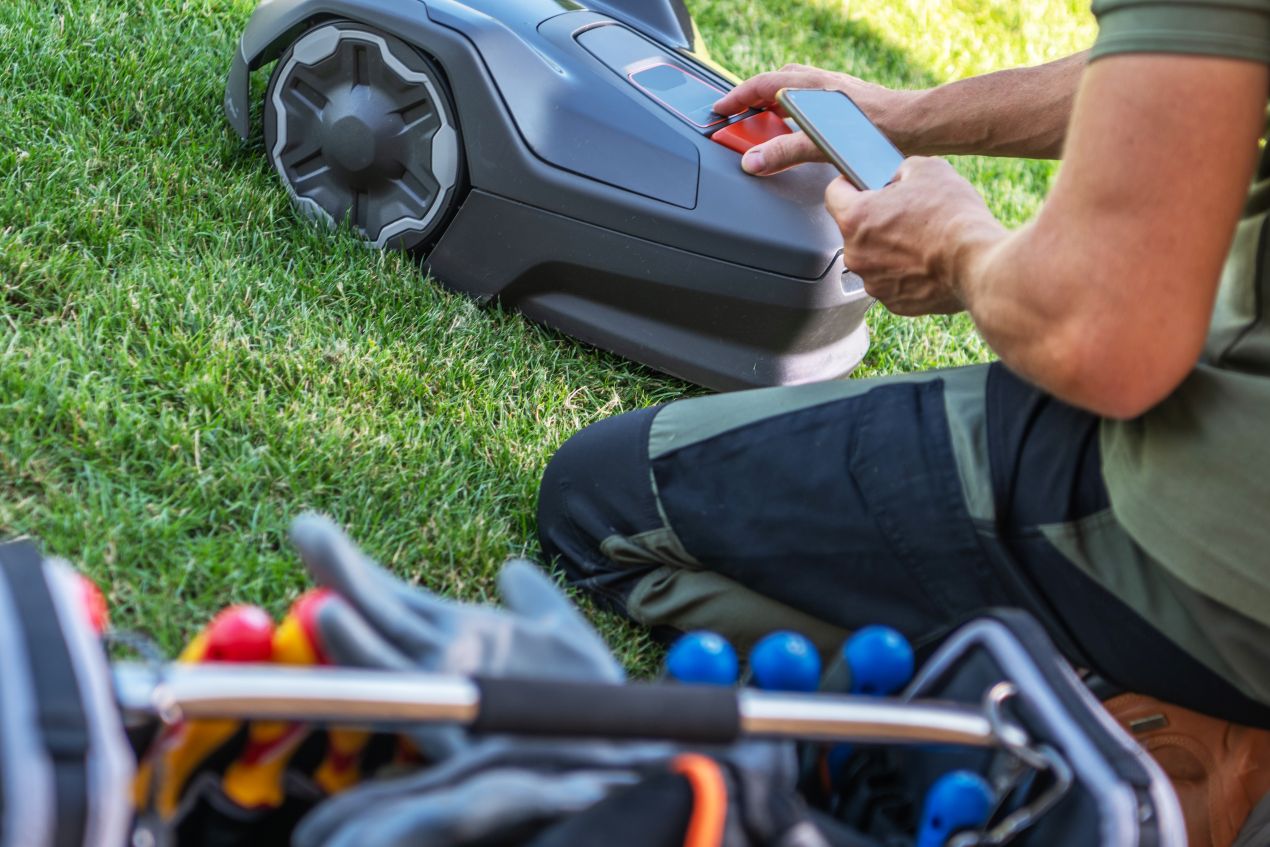 A person configuring a robotic lawn mower using a smartphone while sitting on the grass, with a toolkit nearby.