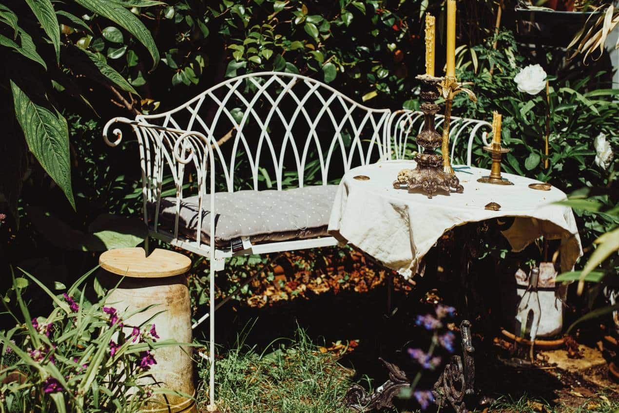 A cozy garden setup featuring a white wrought-iron bench with a gray cushion, placed beside a round table covered with a cloth and brass candle holders, surrounded by lush greenery and flowers. The setting evokes a romantic and nostalgic mood, perfect for reviving love stories in your backyard.