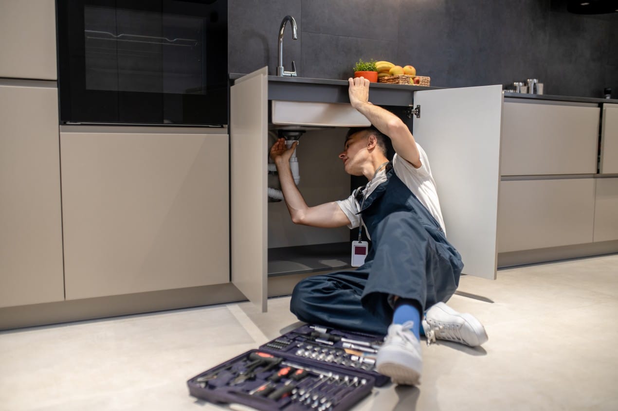 A plumber in overalls is fixing a kitchen sink pipe under a countertop, surrounded by modern cabinets and a toolbox with tools on the floor. Perfect illustration for finding the best plumber in the Southern Highlands.