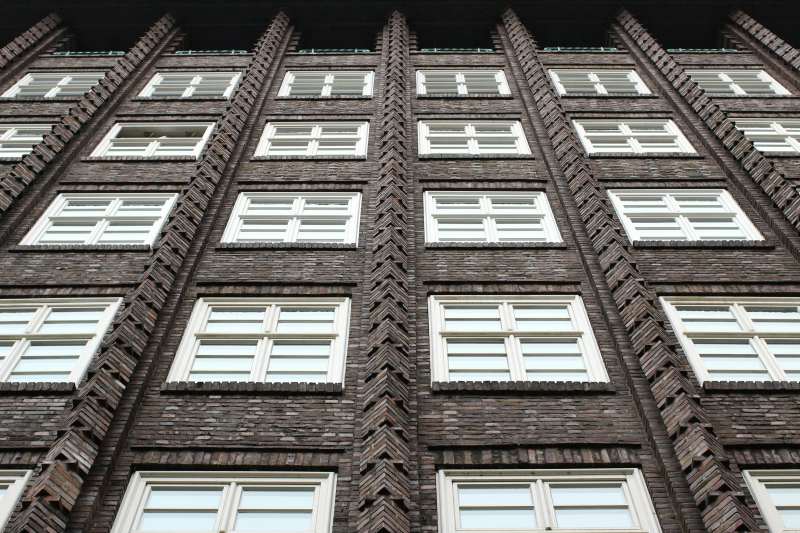 Close-up view of a modern brick building façade with geometric cladding design and multiple white-framed windows arranged in a symmetrical pattern. The cladding highlights texture and depth in construction.