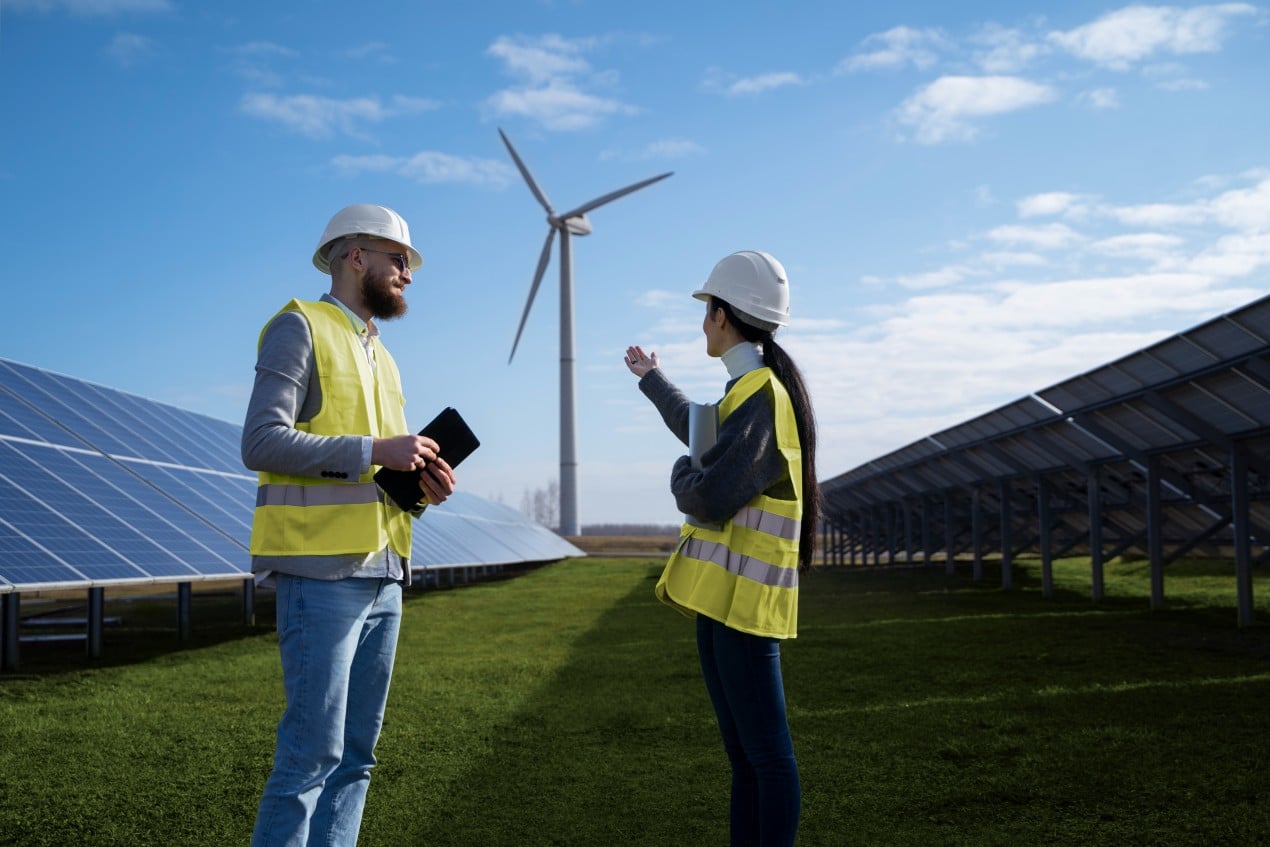 Two engineers wearing safety helmets and reflective vests discuss a project at a renewable energy site with solar panels and a wind turbine in the background, representing clean energy solutions.