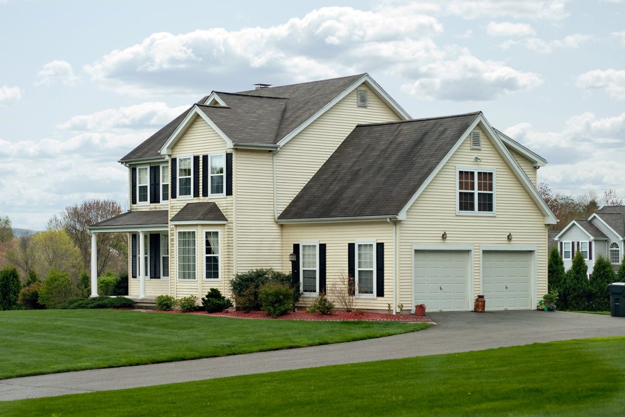 Two-story suburban house with beige siding, black shutters, and a double garage surrounded by a neatly maintained green lawn and driveway.