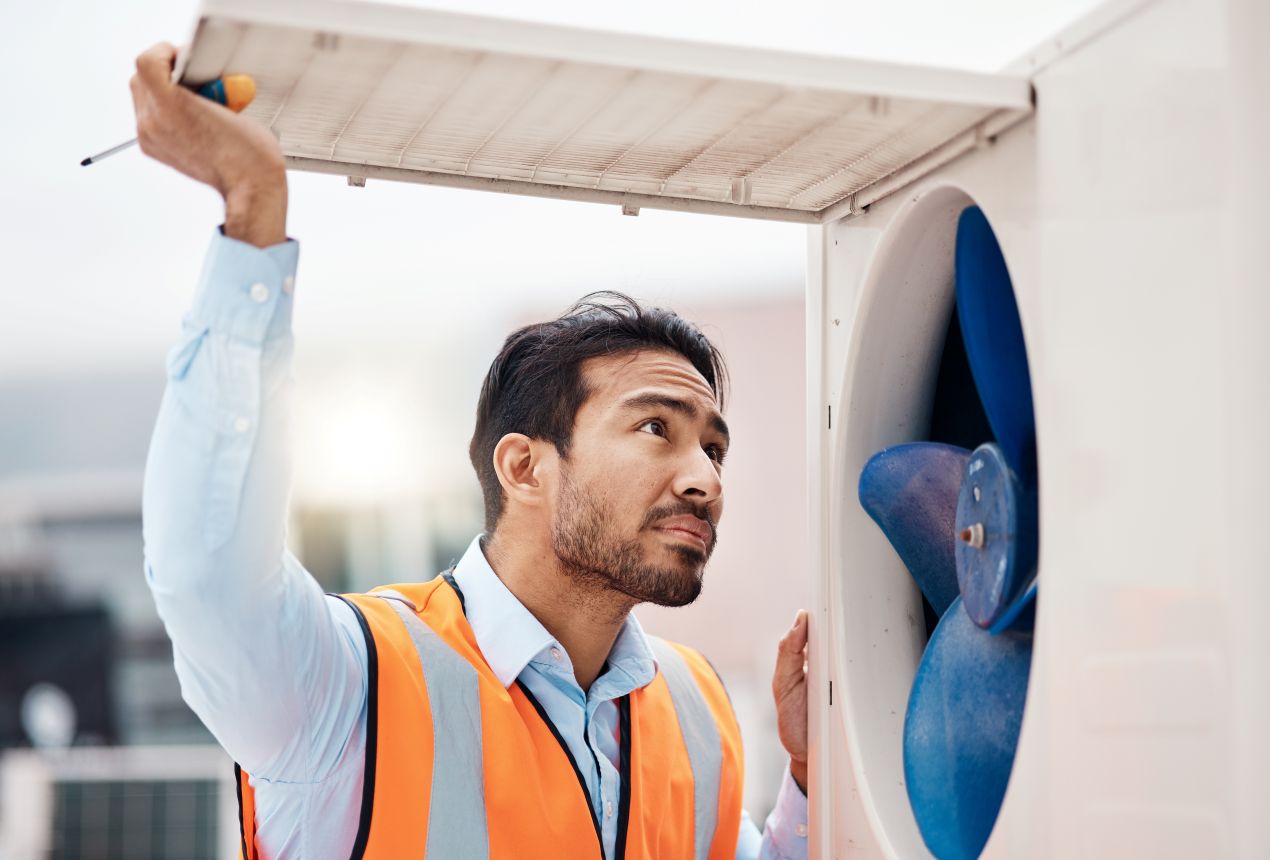 Technician wearing a safety vest inspecting an evaporative cooler unit, checking fan blades and components to ensure efficient air conditioning system performance.