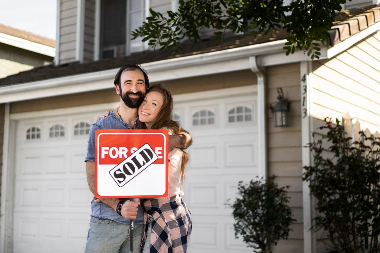 Happy couple standing in front of their new home holding a “For Sale” sign with a “Sold” sticker, symbolizing successful home purchase and avoiding foreclosure.