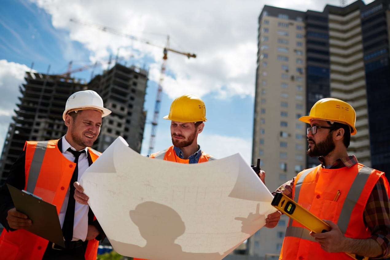 Three construction professionals wearing safety vests and hard hats review blueprints at a building site, with cranes and high-rise structures in the background — illustrating teamwork and planning led by a general contractor for efficient project management.