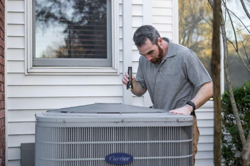 Technician inspecting an outdoor Carrier HVAC unit beside a house, using a flashlight to check components—proven HVAC service in Durant that helps extend system lifespan.