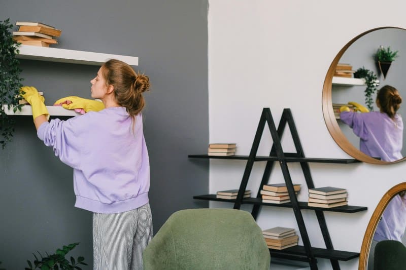 Woman wearing yellow gloves and a lavender sweatshirt cleaning a white shelf in a modern living room with gray and white walls, books, and a round mirror. Represents good home maintenance habits.