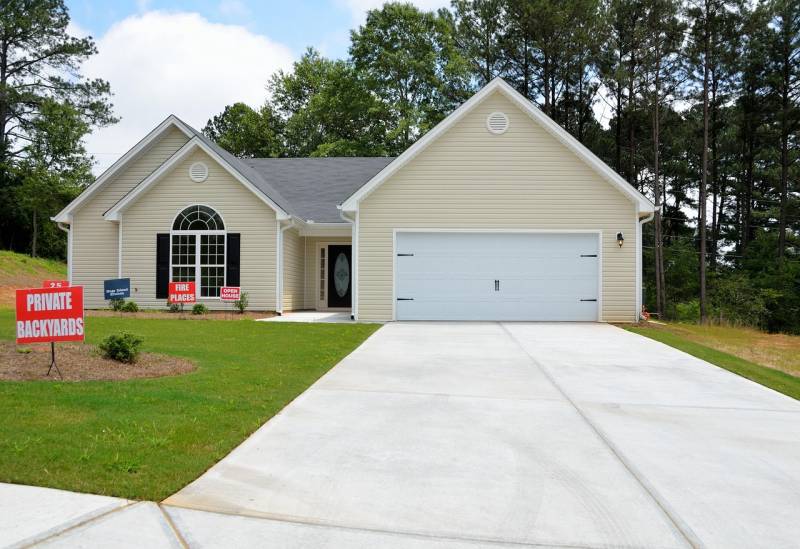 A modern single-story beige house with a two-car garage, wide driveway, and neatly maintained front lawn surrounded by trees. Red signs in the yard advertise features like private backyards and fireplaces, representing a property ready for home selling.