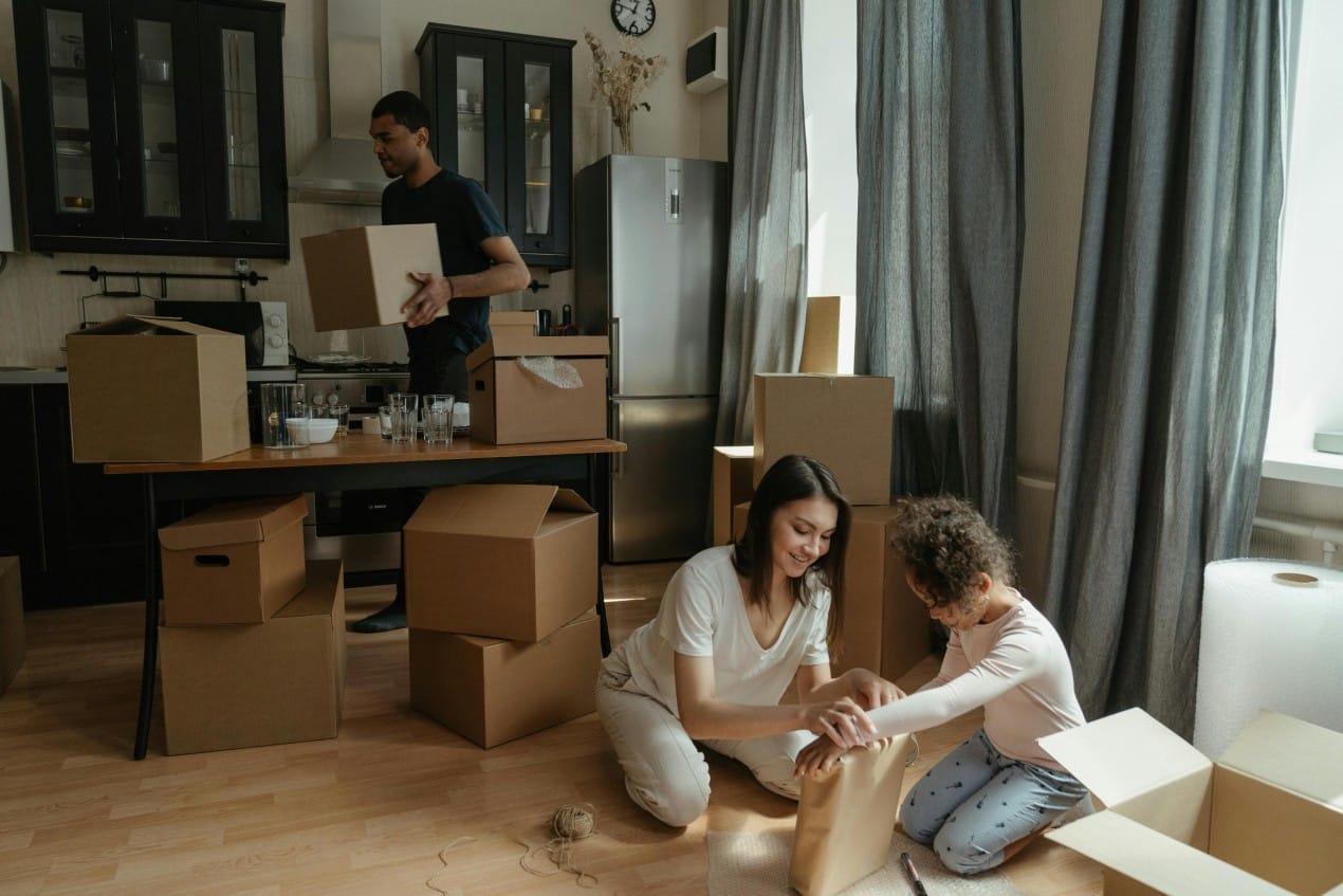 A family is packing boxes in their kitchen, preparing to move into a new home. A man packs items on the table while a woman and child wrap objects on the floor, surrounded by cardboard boxes. The scene captures the start of a new home routine.