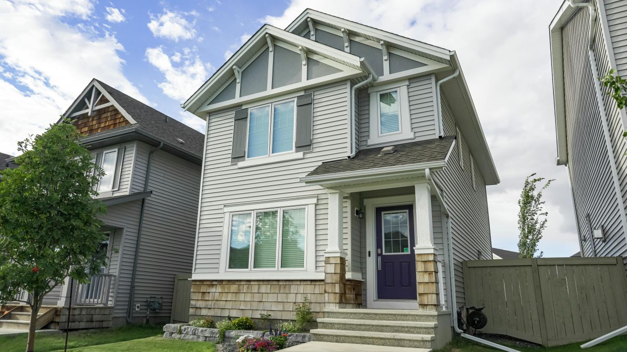 Modern two-story house with gray siding, white trim, and a purple front door. The home’s exterior features decorative gables, stone accents, and a small landscaped front yard with flowers and grass.