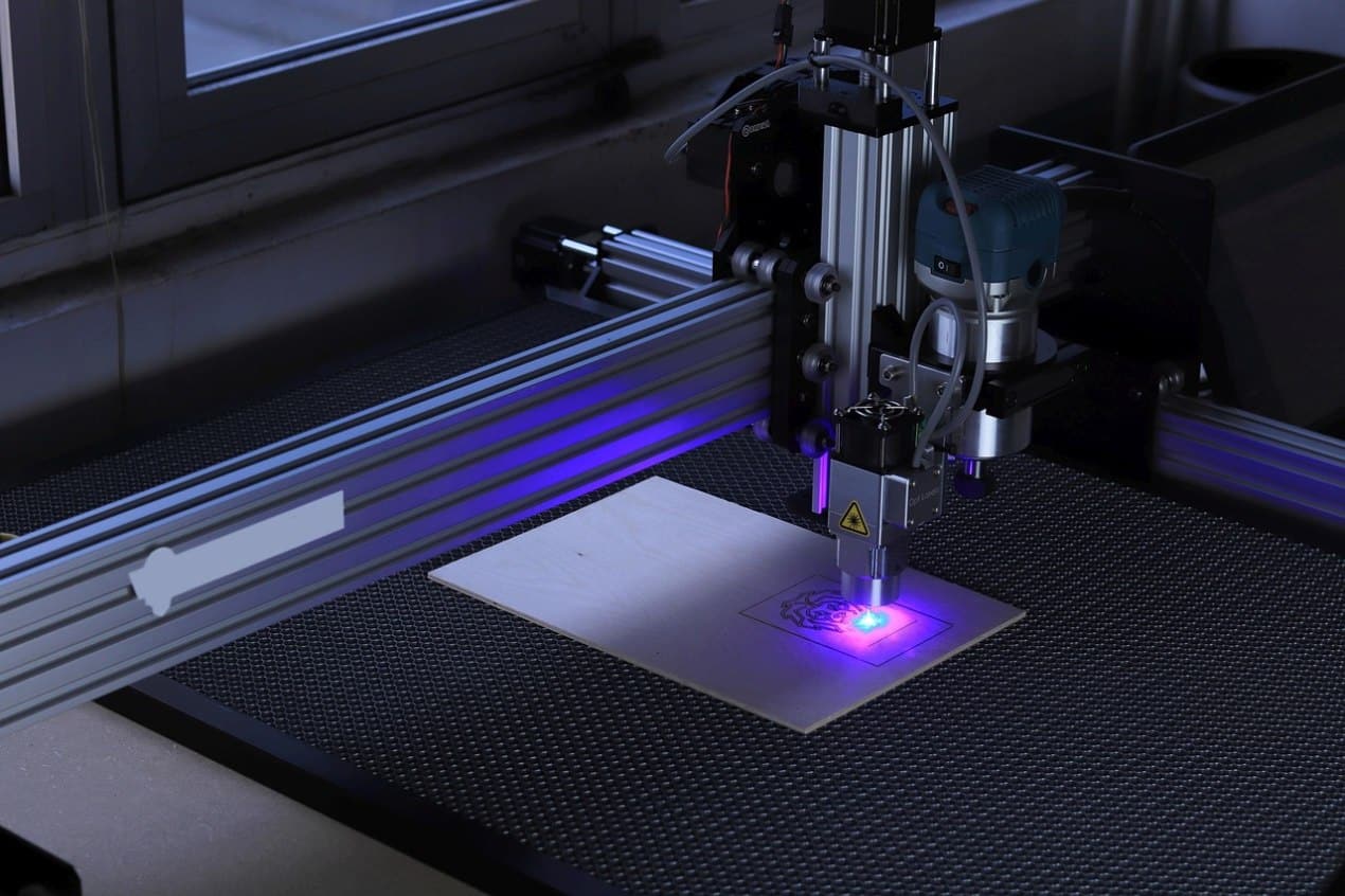 A laser engraving machine in operation etching a design onto a piece of wood, showing precise blue laser light and metal frame components used in interior design and decoration projects.