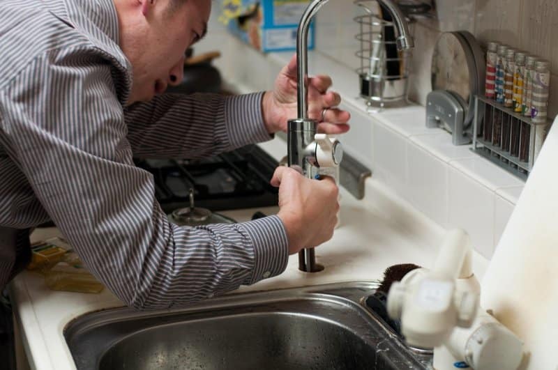 A plumber fixes a kitchen faucet at a sink, using tools to adjust the fitting. The image represents why homeowners should trust local experts for plumbing needs in the suburbs.