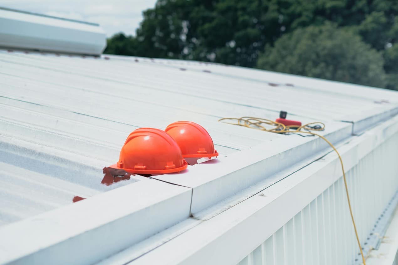 Two orange safety helmets placed on a white metal roof beside a yellow extension cord, symbolizing roofing work and safety practices on a metal roof in Michigan.