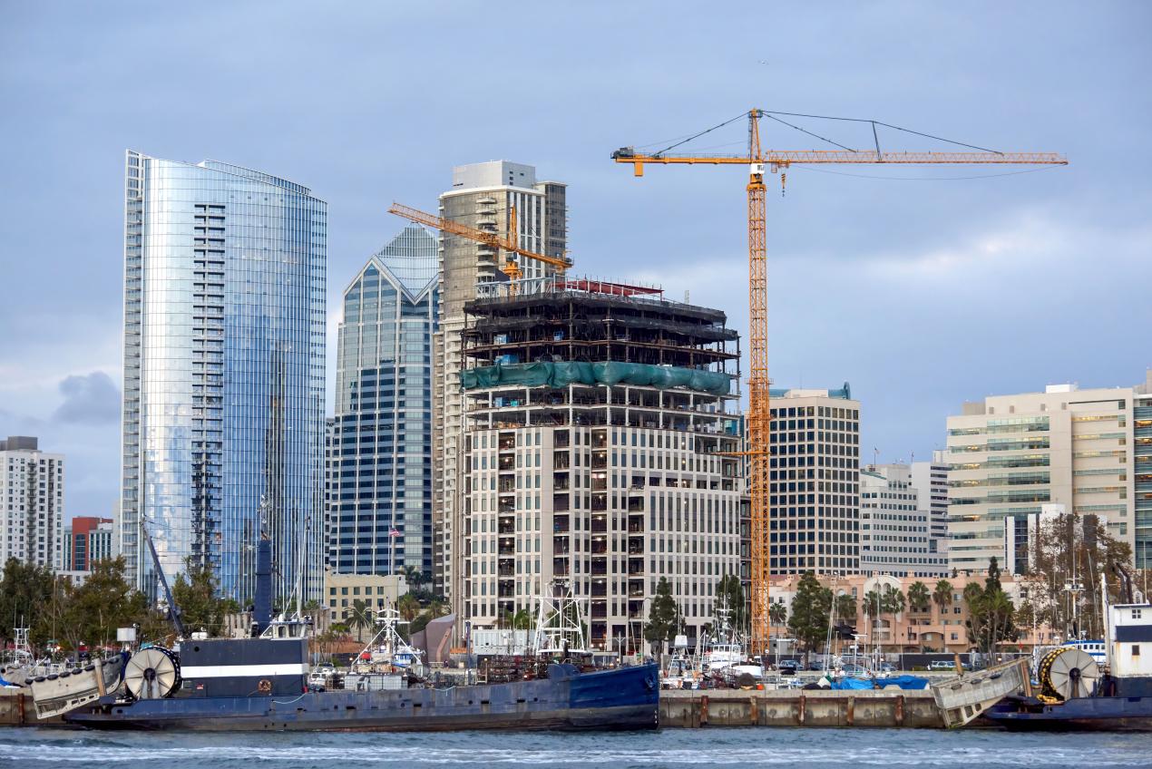 A modern high-rise building under construction with cranes beside it, surrounded by completed glass skyscrapers near the waterfront; an example of a durable building structure being built to last.