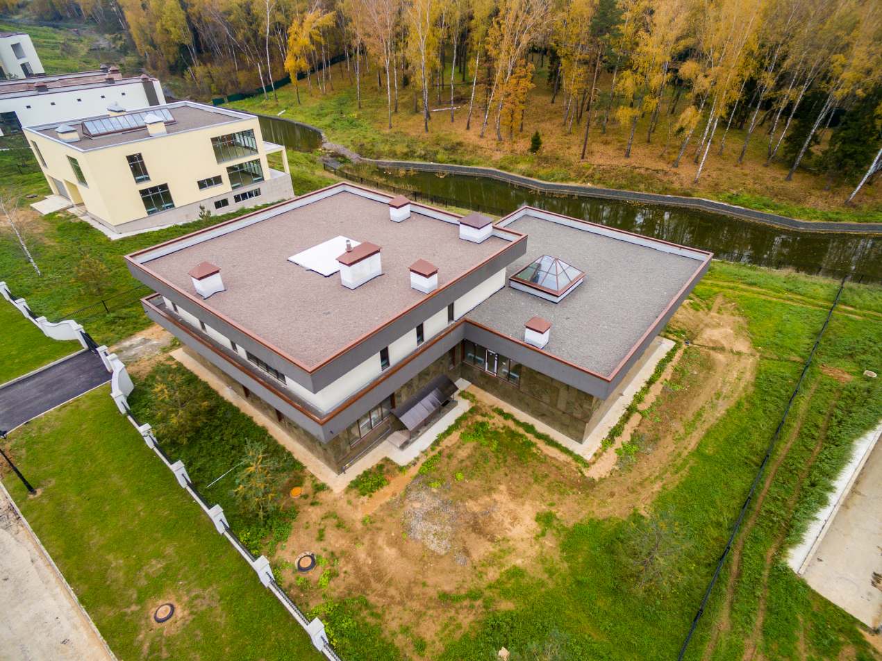 Aerial view of a modern flat-roofed house near a small canal, surrounded by grass and trees, showcasing the clean architectural lines and practical benefits of flat roofing.
