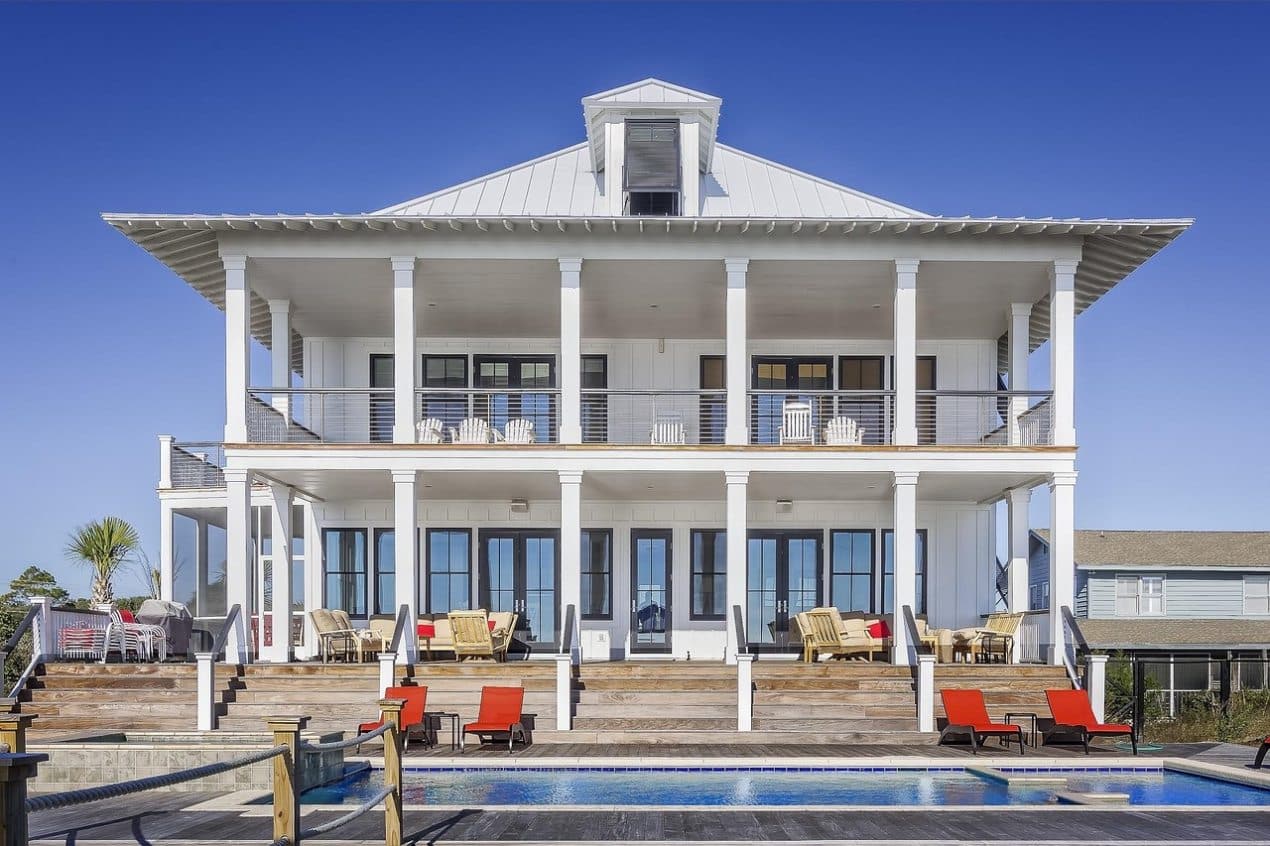 Two-story modern beach house with large white columns, spacious balconies, glass doors, outdoor seating areas, and a swimming pool in front under clear blue sky. Ideal house and land package for luxury living.