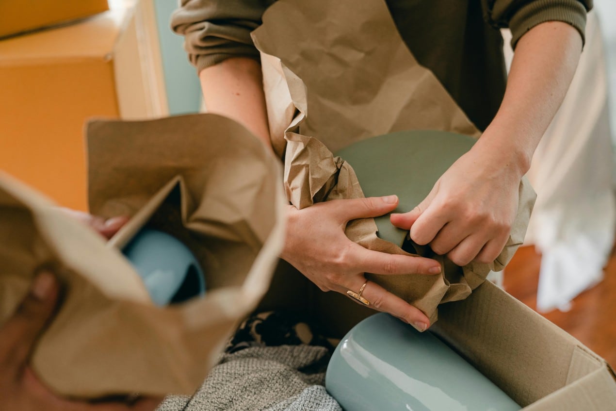 Person carefully wrapping ceramic dishes in brown packing paper while packing boxes, showing the importance of planning ahead when moving.