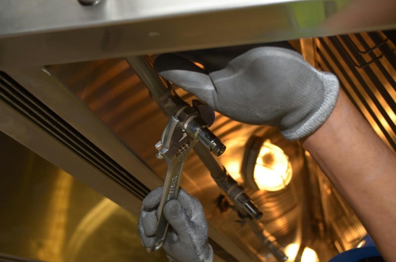 A plumber wearing gray gloves uses a wrench to tighten a metal pipe connection under a stainless steel fixture. The scene is well-lit, showing a close-up of precise plumbing repair work.