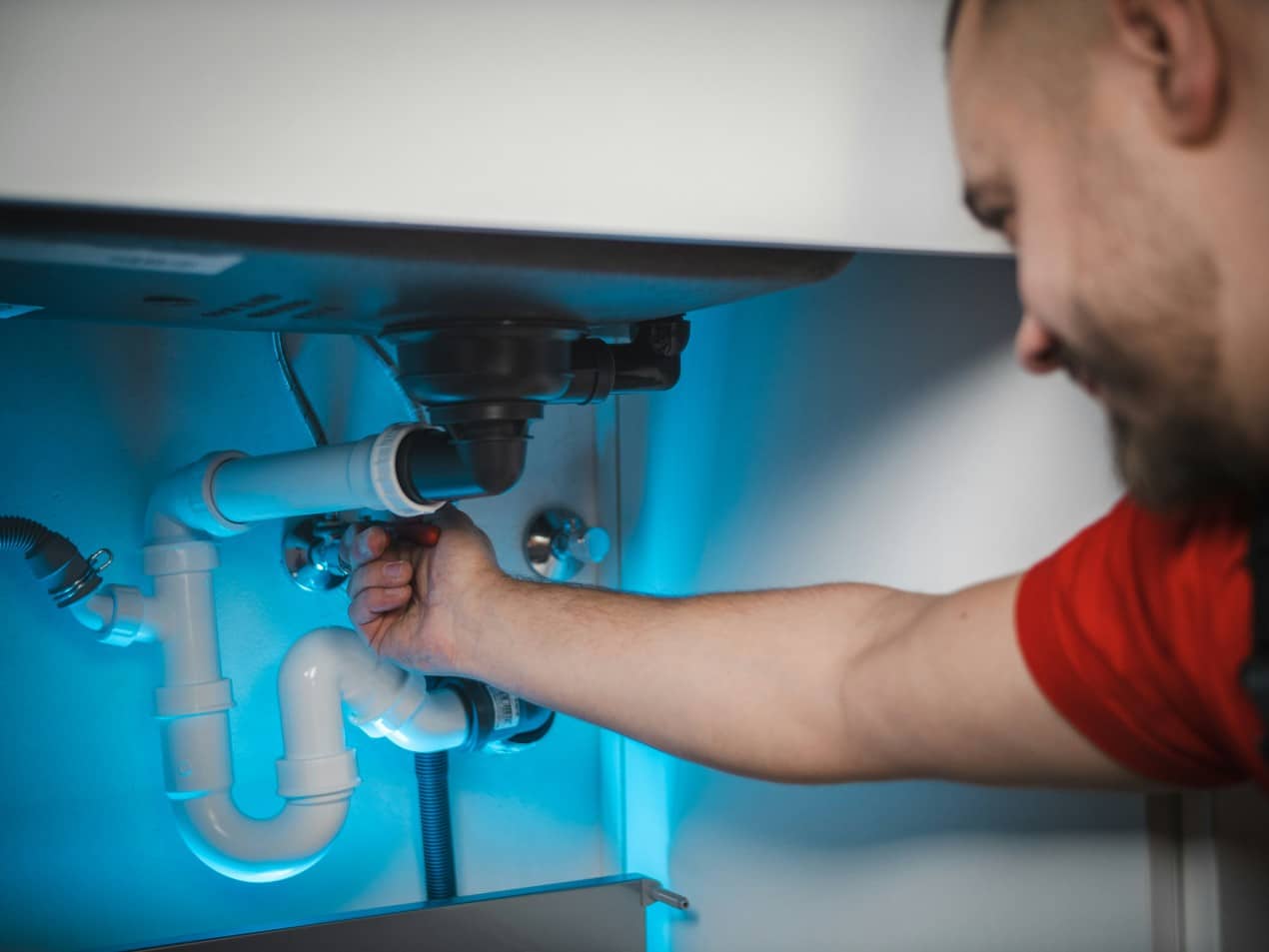 A plumber working under a kitchen sink, tightening a white PVC pipe connection as part of a plumbing system upgrade.