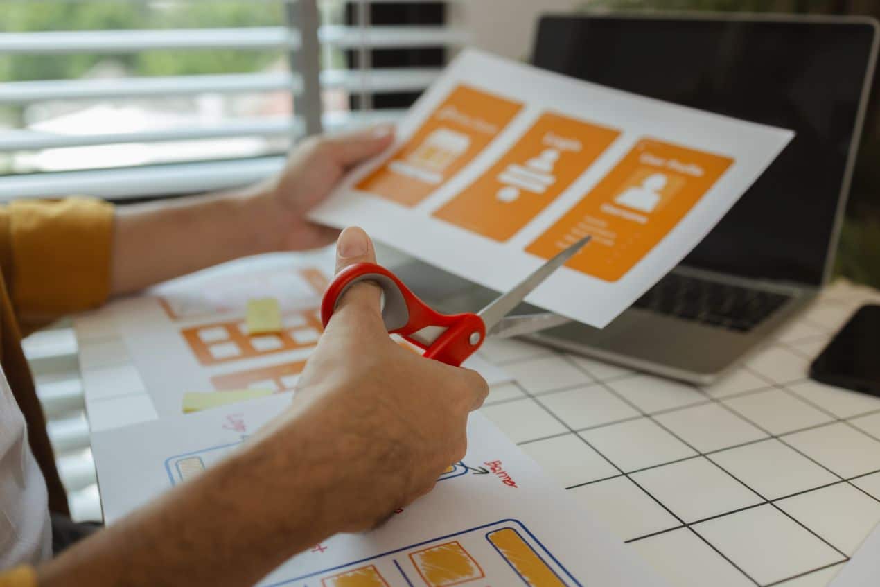 Designer cutting printed layouts with red scissors beside a laptop while refining print materials to ensure consistent brand design and visual alignment.