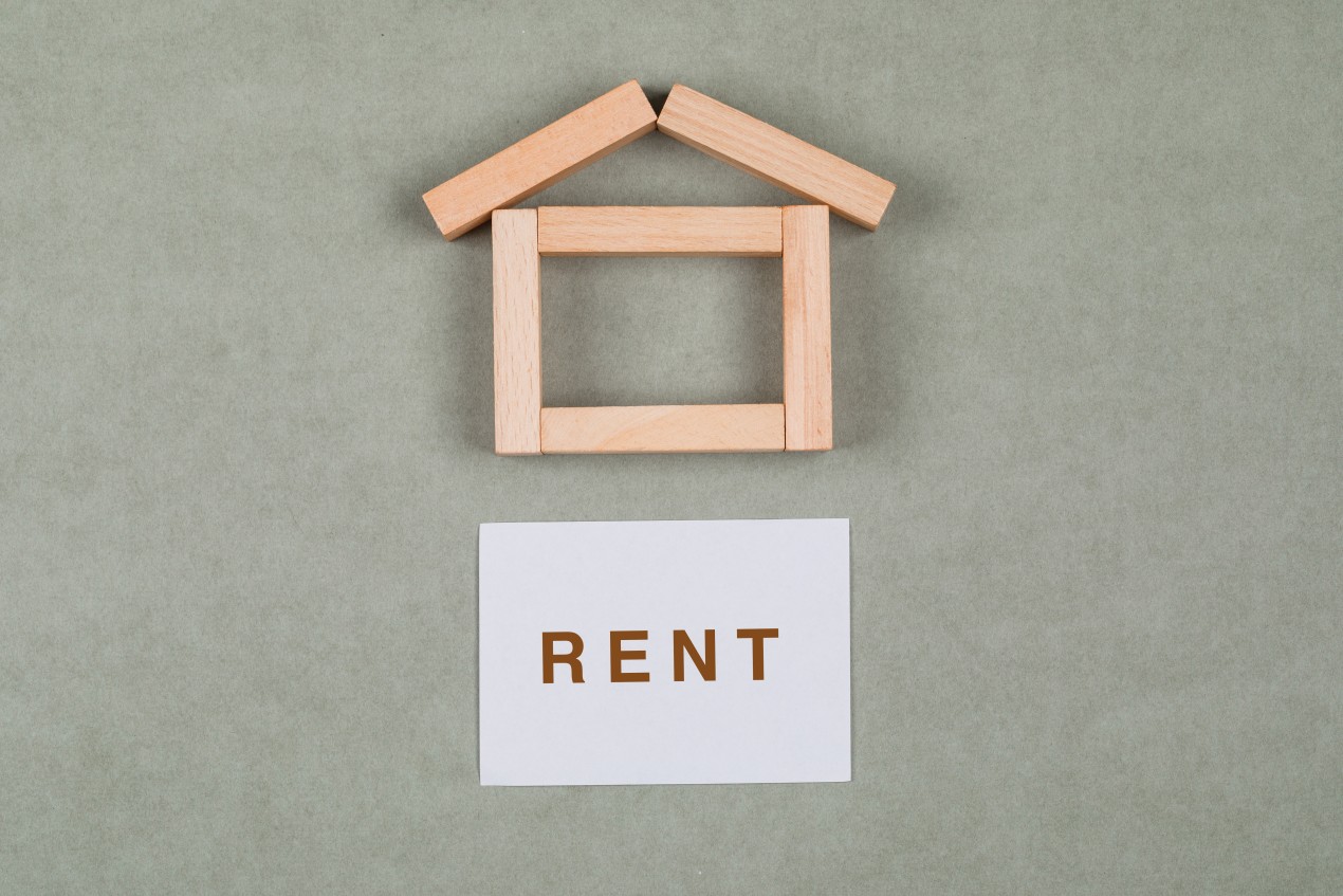 Wooden toy blocks arranged in the shape of a house with a white card below that reads "RENT," representing renting a house or apartment abroad.