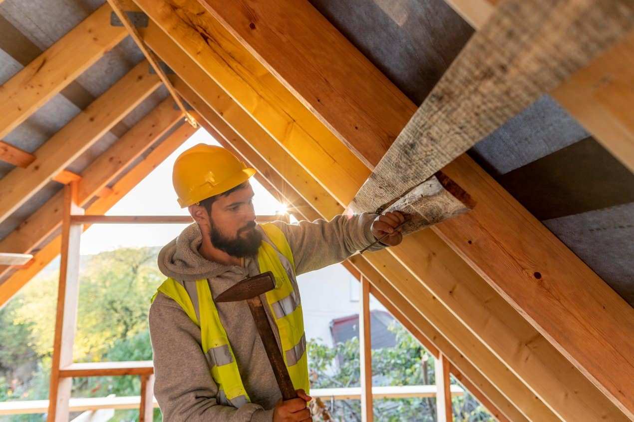 A skilled construction worker wearing a yellow hard hat and safety vest is installing wooden panels inside a roof structure, using a hammer to secure the beams during roof repair.