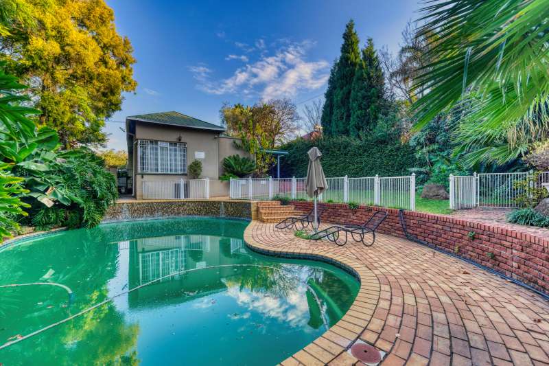 Backyard with a curved swimming pool surrounded by brick paving, greenery, and a safety fence near a house. The swimming pool reflects the sky and trees, creating a calm outdoor setting.
