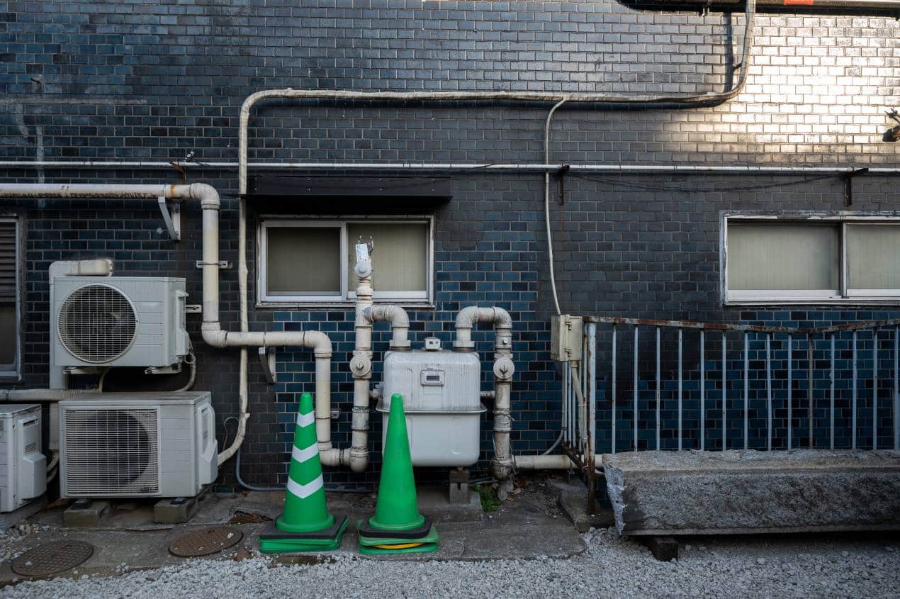 Exterior wall with air conditioning units, exposed pipes, and two green traffic cones near a water or gas meter; an example setup relevant to water heat pump systems.