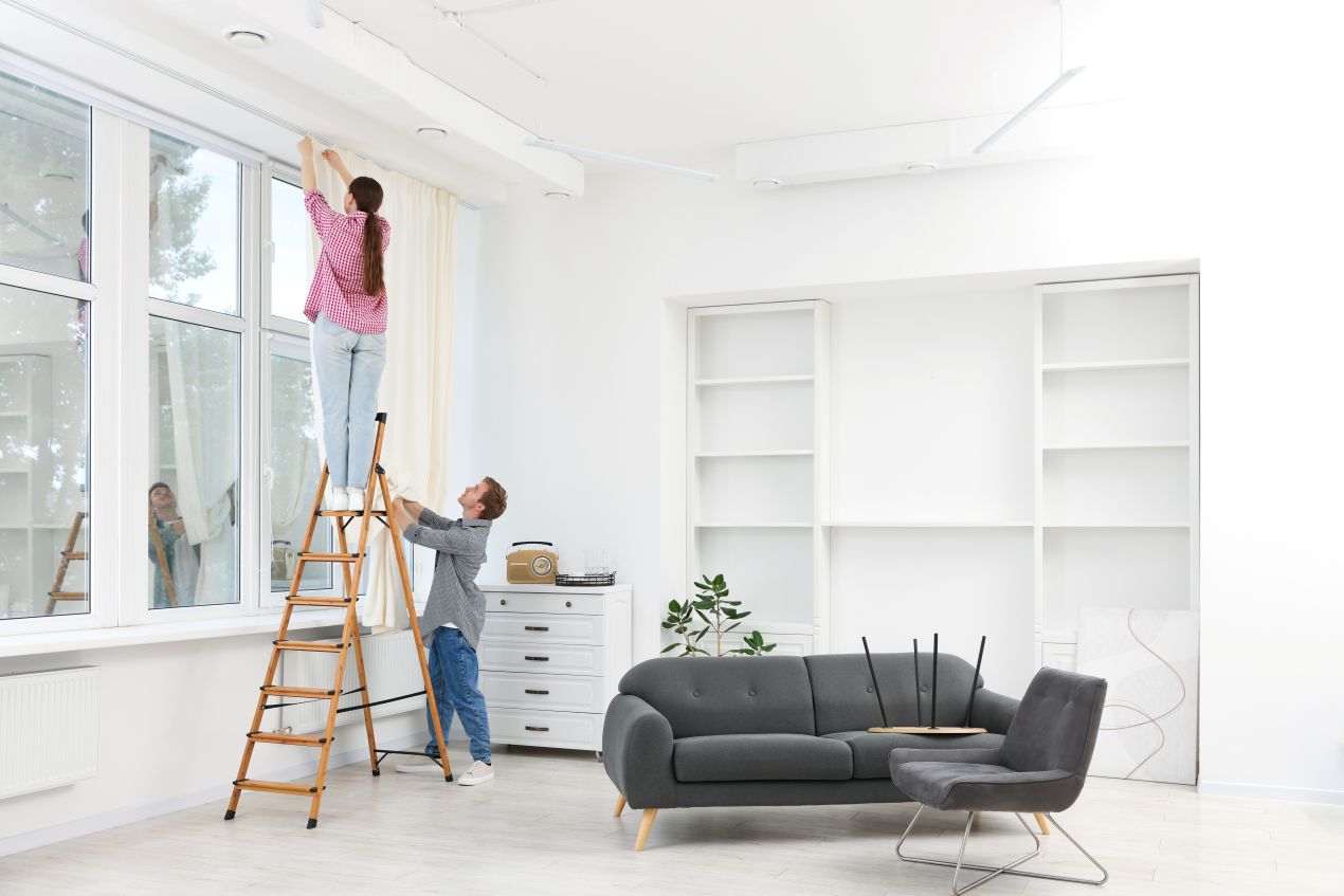 Couple hanging long curtains in a bright living room with minimalist décor and large windows.