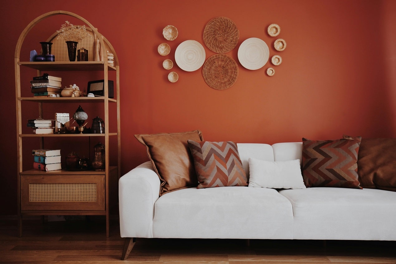 Cozy living room with a terracotta accent wall, white sofa, warm earth tone cushions, rattan wall decor, and wooden shelving showcasing current painting trends.