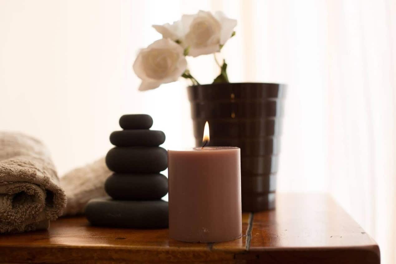 Lit candle on a wooden table with stacked black spa stones, folded towels, and white flowers in a vase, creating a calm wellness-inspired interior design setting.
