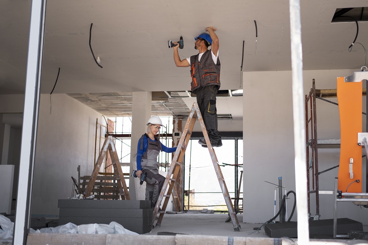 Two home renovation contractors installing ceiling wiring and drywall, one standing on a ladder while drilling overhead