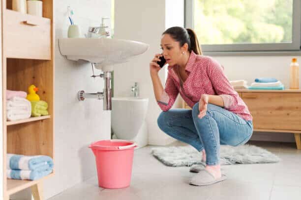 Woman experiencing a plumbing emergency crouches by a leaking bathroom sink with a bucket, calling a plumber for help
