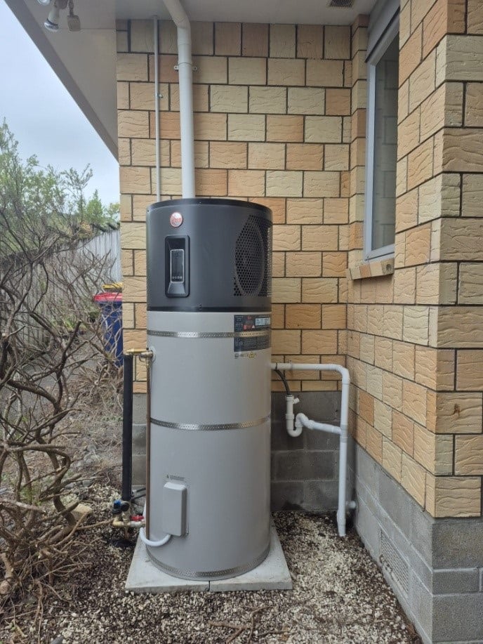 Heat pump hot water system installed outside a brick home, showing the compressor unit and tank